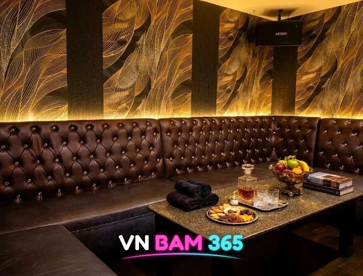 다낭 넘버원 가라오케 VIP 룸, 금빛 깃털 무늬 벽과 갈색 가죽 소파, 위스키 디캔터와 과일이 놓인 테이블
