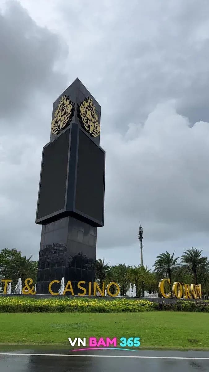 푸꾸옥 코로나 리조트 앤 카지노 도로변의 금색 '& CASINO CORONA' 글자 조형물과 금색 문장이 달린 검은 사각 타워, 뒤편 분수와 야자수